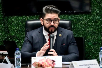Un hombre barbudo con gafas y traje oscuro, camisa blanca y corbata roja, habla en un micrófono mientras gesticula con la mano en una mesa