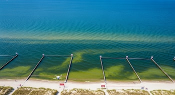 Vista aérea de la costa del mar Báltico con agua azul y manchas verdes, arena, playas delimitadas por cintas rojas, carteles, espigones de madera y un dron.