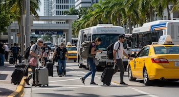 Personas con maletas y mochilas caminan por una calle con taxis amarillos y autobuses en Miami. Al fondo, edificios, palmeras y un letrero de 'Brightline'.