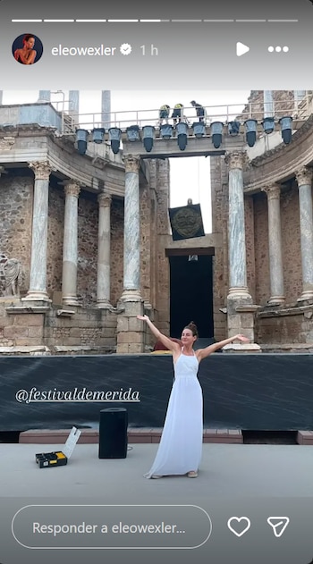 Una mujer de vestido blanco con los brazos abiertos en un escenario. Al fondo, un teatro romano con columnas. Hay luces, un altavoz y una caja de herramientas en el suelo