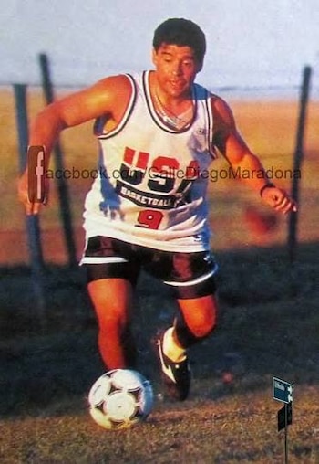 Un hombre (Diego Maradona) con camiseta blanca de baloncesto con la leyenda "USA BASKETBALL 9" y pantalón corto negro, dribla un balón de fútbol blanco y negro en un campo de césped