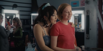 Poppy Liu, con cabello oscuro y top de mezclilla, abraza a Hannah Einbinder, de cabello rubio y suéter rojo, en un camerino