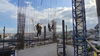 Tres obreros con cascos y arneses están sobre una estructura de varillas de acero. Una grúa y edificios se observan al fondo con cielo nublado