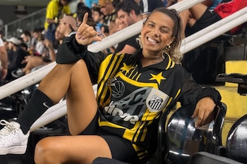 Mujer sonriente sentada en la tribuna de un estadio con una camiseta negra y amarilla con el escudo del Santos FC y calzado deportivo blanco