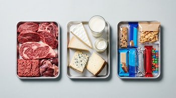 Tres bandejas metálicas con carne roja (cortes y molida, vísceras), productos lácteos (quesos variados, leche, yogur) y alimentos ultraprocesados empaquetados.