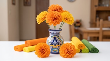 Flores naranjas y amarillas de papel en un florero de cerámica decorado, sobre una mesa blanca junto a rollos de papel crepé.