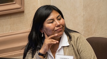 Mujer de cabello oscuro con chaqueta beige y camisa blanca que apoya la mano en la cara frente a una tarjeta con la palabra conferencia.