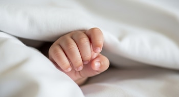 Primer plano de una mano de bebé con los dedos flexionados que sale de una sábana o manta blanca.