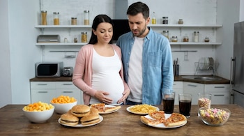 Una mujer embarazada y un hombre de pie frente a una mesa con hamburguesas, patatas fritas, pizza, refrescos, snacks de queso y cereales de colores.