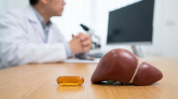 Una cápsula de gelatina blanda naranja y un modelo anatómico de hígado marrón rojizo sobre una mesa de madera. Un profesional médico borroso y equipo de laboratorio al fondo.