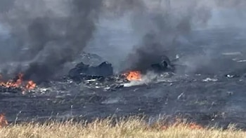 Llamas y humo negro se elevan desde restos carbonizados sobre un terreno quemado, con pastos secos en primer plano