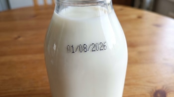 Botella de vidrio transparente con leche blanca y la fecha 01/08/2026 impresa, sobre una superficie de madera.