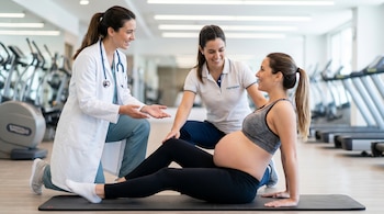 Una mujer embarazada sentada en un tapete de ejercicio es asistida por una médica y una fisioterapeuta en un gimnasio.