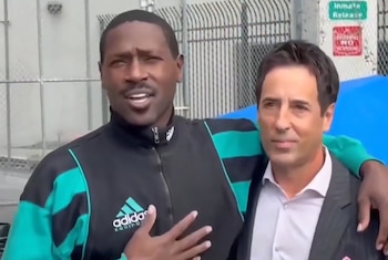 Antonio Brown viste una chaqueta deportiva negra y verde mientras abraza por el hombro a un hombre en traje frente a una cerca metálica