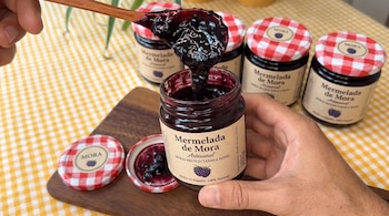 Manos sujetan un frasco de mermelada de mora y una cuchara de madera frente a otros frascos sobre un mantel a cuadros.