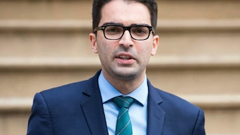 Hombre con gafas de montura negra, saco azul marino, camisa celeste y corbata verde a rayas frente a una escalera de piedra