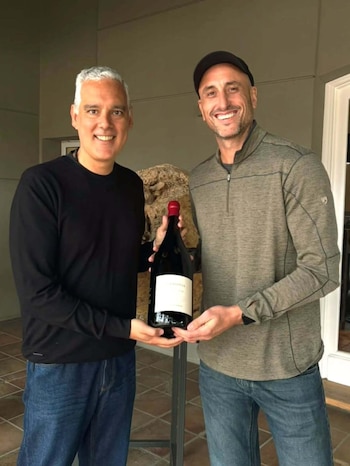 Dos hombres de pie sostienen una botella de vino de gran formato en una galería con una escultura de piedra al fondo