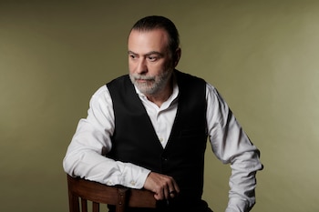 Reynaldo Sietecase, hombre con barba gris, cabello recogido, camisa blanca y chaleco negro, sentado en una silla de madera, mirando a la izquierda
