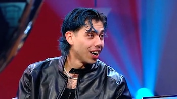 Luck Ra, un joven con cabello oscuro y chaqueta de cuero negra, sentado frente a un entrevistador de traje gris en un set de televisión con luces azules