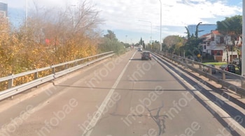 Un puente de asfalto con barreras metálicas, árboles, casas a la derecha, postes de luz y automóviles en circulación.