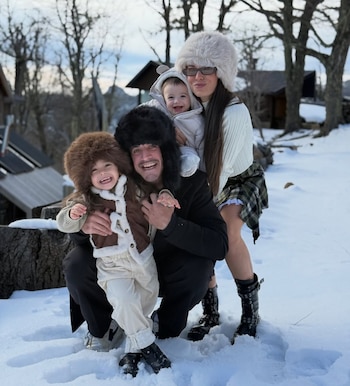China Ansa y Diego Mendoza con sus dos hijos pequeños sonríen y posan en la nieve, vistiendo ropa de invierno y sombreros de piel. Al fondo hay árboles y cabañas de madera