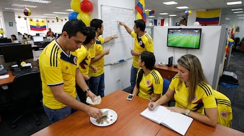 Grupo de personas con camisetas de la selección de Colombia en una oficina: un hombre cuenta billetes y monedas, una mujer escribe en una agenda, y otros miran un marcador.