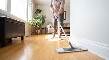 Una persona adulta de pie usa una mopa plana rectangular para limpiar un zócalo blanco en una sala de estar con piso de madera clara junto a una ventana.