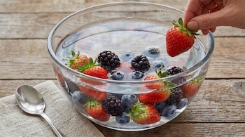 Primer plano de una mano introduciendo una fresa en un cuenco de cristal transparente con agua, fresas, arándanos y moras. El cuenco está sobre una mesa de madera.