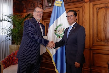 Dos hombres con traje formal se dan la mano de pie junto a la bandera de Guatemala en una sala con paredes de madera