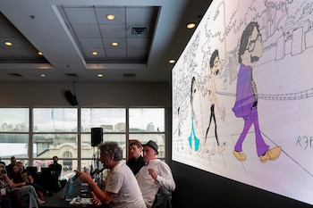 Fito Páez, hombre de pelo rizado, habla en un evento con micrófono. Detrás, una pantalla LED muestra una caricatura de un hombre con gafas y traje morado
