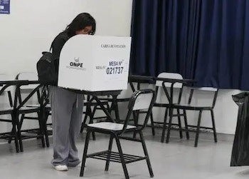 Mujer joven con camiseta negra y pantalón gris votando dentro de una cabina electoral blanca de ONPE, en un aula con mesas y sillas. Fondo con cortina azul oscura