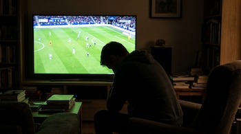 Silueta de una persona de espaldas y con la cabeza gacha, sentada en un sillón oscuro frente a un televisor que muestra un partido de fútbol en un salón tenue.