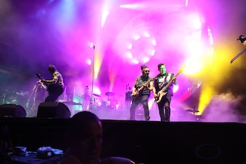 Tres músicos, incluyendo dos guitarristas y un bajista, actúan en un escenario con luces de color púrpura y amarillo y humo. Batería visible al fondo