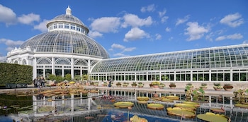 Gran Conservatorio Enid A. Haupt, con cúpula y paredes de cristal, en el NYBG, frente a un estanque con nenúfares grandes y el cielo azul con nubes blancas