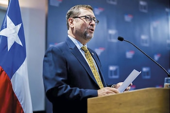 Primer plano de un hombre con barba y gafas, vestido con traje y corbata, hablando en un podio con documentos. A su izquierda se ve parte de la bandera chilena
