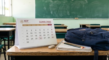 Calendario de la SEP de junio de 2026 con el 26 marcado, sobre un pupitre de madera junto a una mochila azul, cuadernos, lápices y un borrador en un aula.