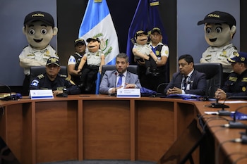 Autoridades sentadas en una mesa frente a micrófonos con marionetas y botargas de la policía detrás de ellas y la bandera de Guatemala