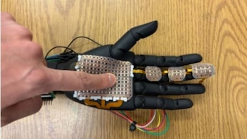 Un dedo humano toca un módulo sensor flexible sobre la palma de una mano robótica negra colocada en una superficie de madera