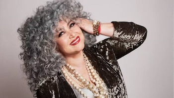 Primer plano de Amanda Miguel sonriendo, con cabello gris rizado, labios rojos, múltiples collares de perlas y una chaqueta oscura de terciopelo