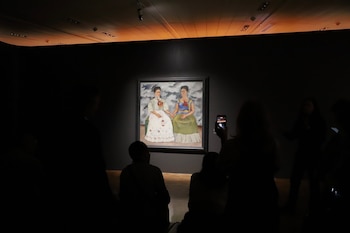 Pintura con dos figuras femeninas sentadas y tomadas de la mano, ambas con corazones expuestos, exhibida en sala oscura con público
