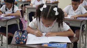 Una niña con lazos blancos en el cabello dibuja en un cuaderno sobre su escritorio en un salón de clases con otros alumnos.
