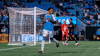 Jugador de fútbol con camiseta celeste y pantalón blanco número 25 corriendo en campo de césped. Hay un arco, otros jugadores y público en el estadio.
