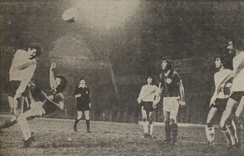 Fotografía en blanco y negro de un partido de fútbol, mostrando a un jugador realizando una patada aérea mientras la pelota está en el aire, con otros jugadores y un árbitro en el campo