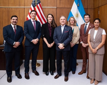 Siete personas con traje formal posan de pie delante de las banderas de Estados Unidos y Argentina sobre una pared de madera