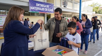 Mujer entrega bolsa con "KIT ESCOLAR" a hombre. Niño observa útiles en caja. Fila de personas detrás. Un cartel azul dice "ENTREGA DE MATERIALES DE CLASE".
