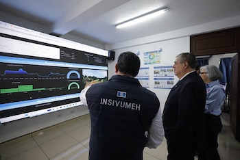 Tres personas observan una pantalla grande que muestra gráficos de datos hidrológicos y una imagen de paisaje, con posters informativos en la pared del fondo