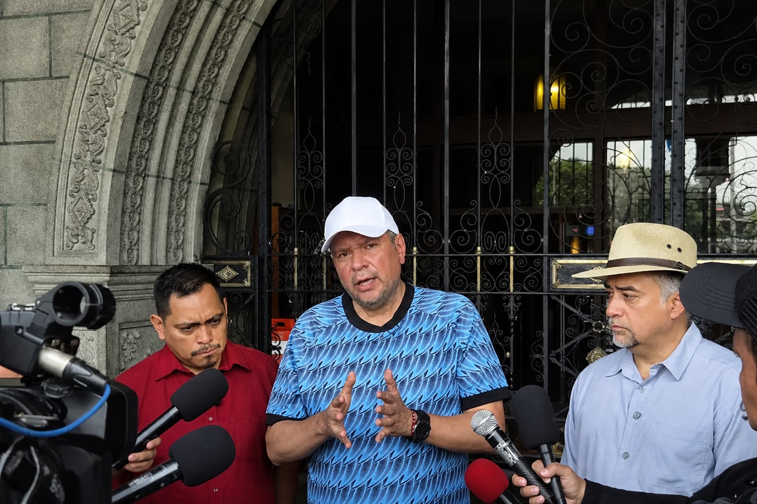 Un hombre con gorra blanca realiza declaraciones ante los medios de comunicación rodeado de periodistas y grabadoras frente a un edificio. (Imagen Ilustrativa Infobae)