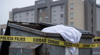 Contenedor de basura metálico con una sábana blanca, cinta policial amarilla FGJES alrededor, frente a un edificio de hotel.