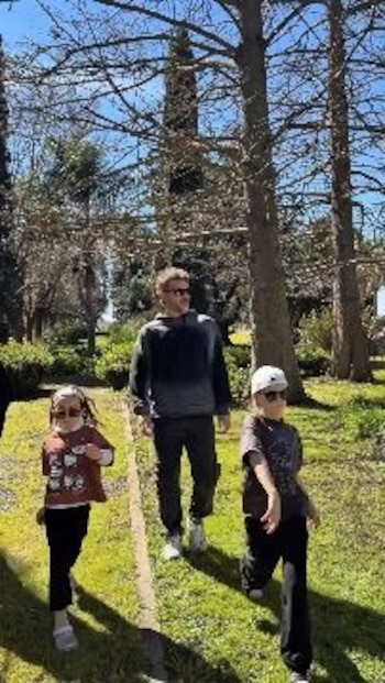 Un hombre y dos niños con gafas de sol caminan sobre el césped de un jardín rodeado de árboles