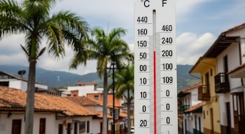 Primer plano de un termómetro blanco con mercurio rojo que indica alta temperatura, con palmeras, techos de teja y montañas al fondo.